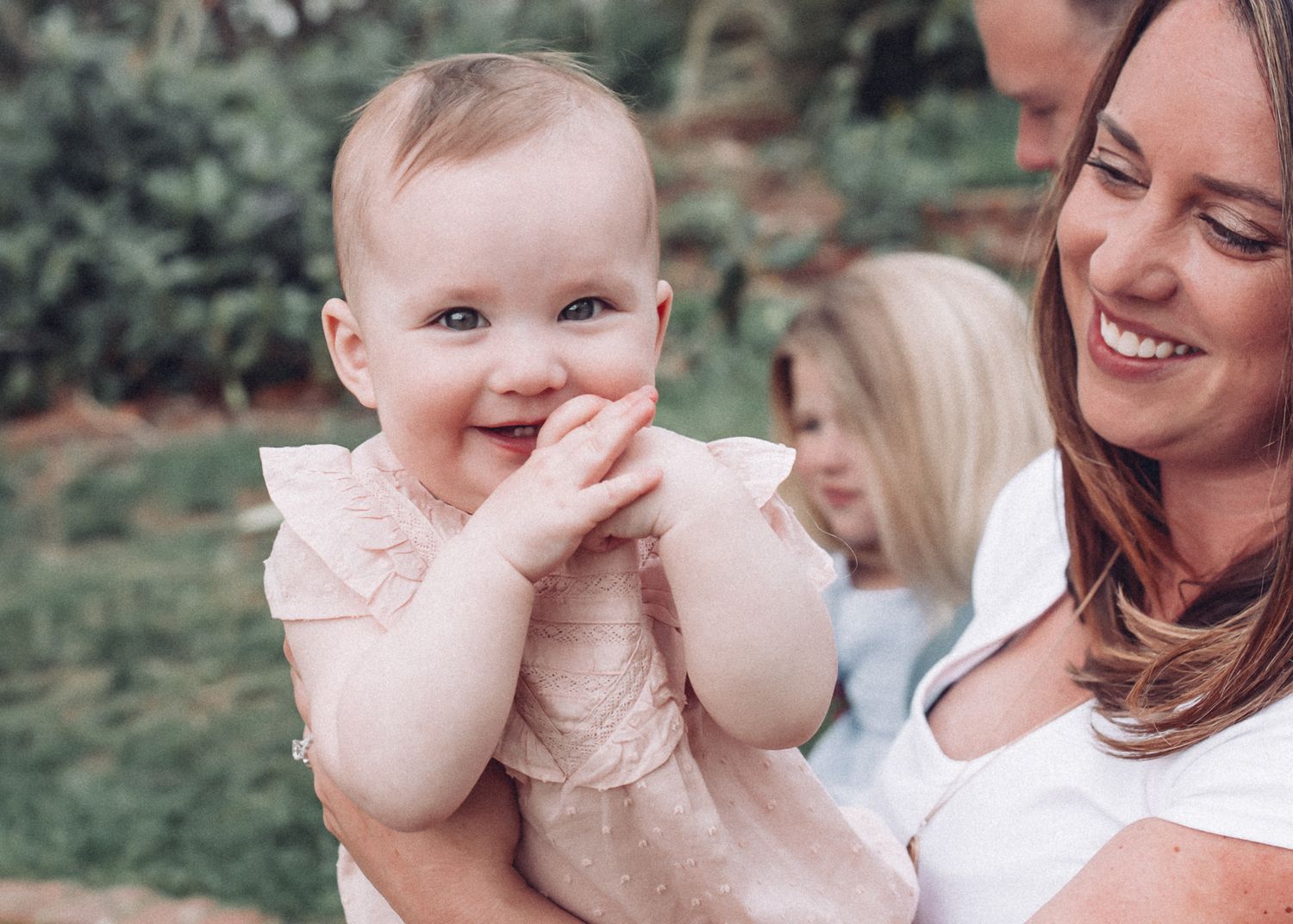 A baby in a ruffled dress giggles with hands near face while being held outdoors.