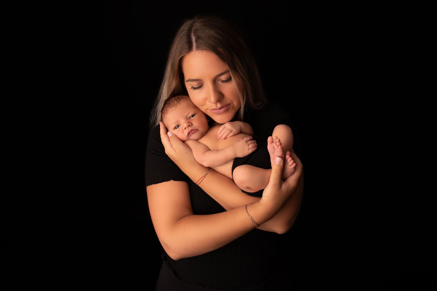 A mother in black holds her newborn baby tenderly against a dark background in an intimate portrait session.
