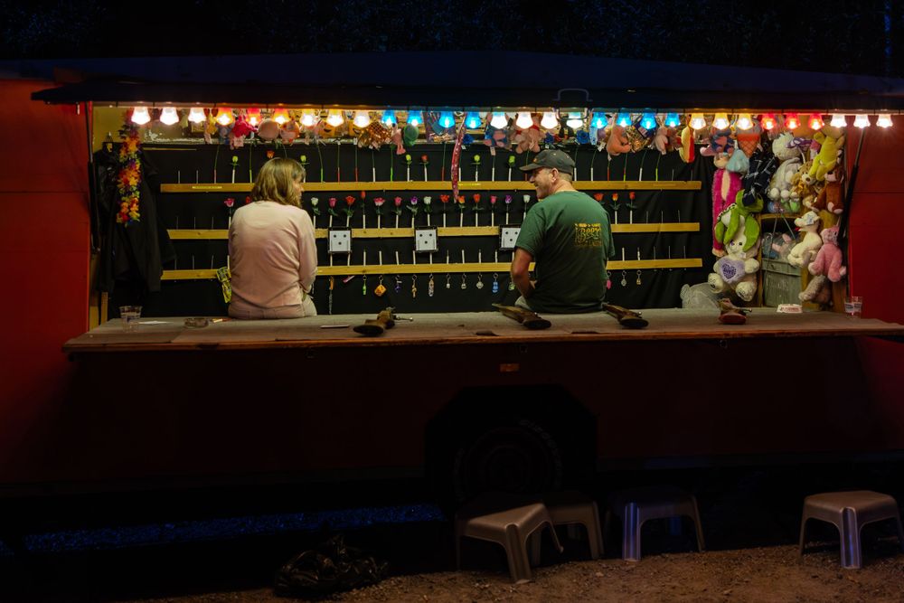 Two patrons sit at a dimly lit bar with string lights overhead and multiple bottles displayed on shelves.