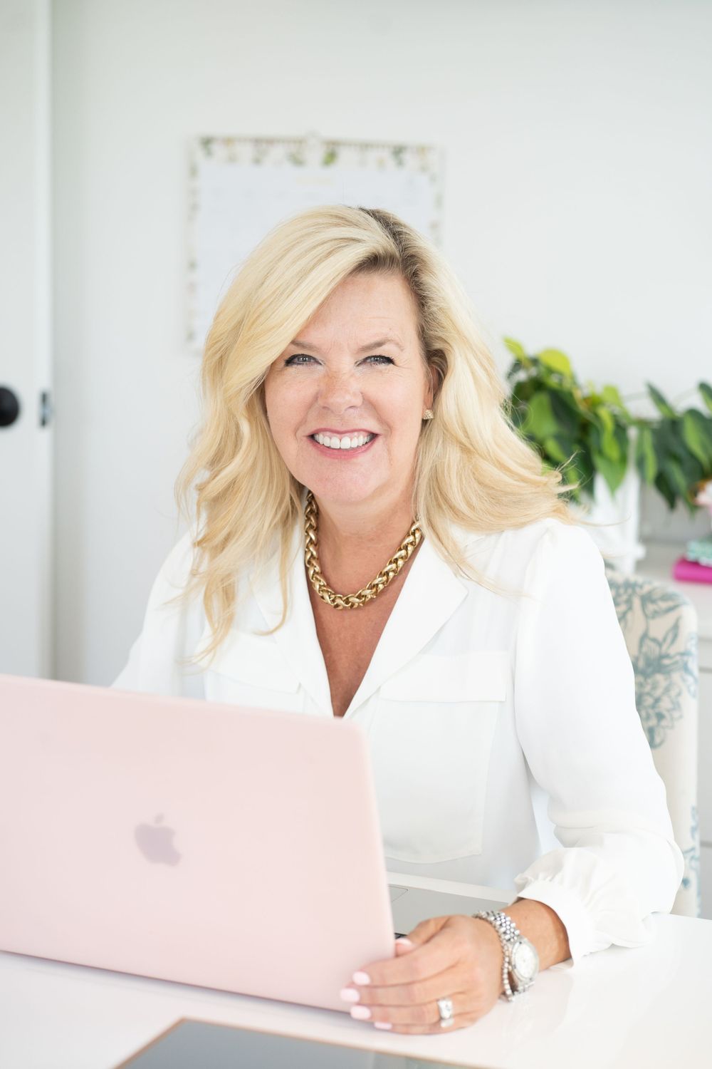 Professional woman in white blazer working on pink laptop at desk.
