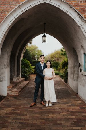Elegant couple poses under arched brick passageway with hanging lantern and garden views.