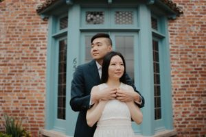 Intimate embrace against vintage brick building with teal bay window architectural details.