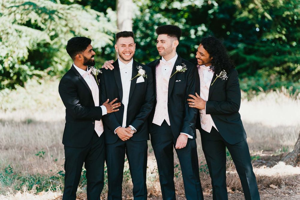 Four groomsmen in black tuxedos and white bowties share a candid laughing moment outdoors.