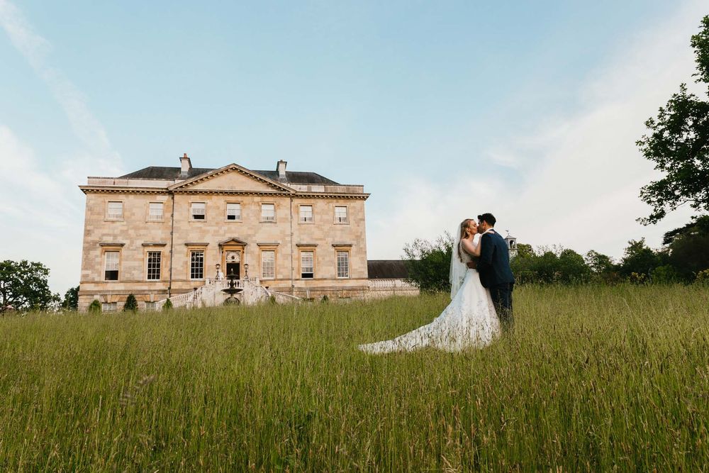 Newlyweds pose in front of a grand historic mansion on a grassy hillside.