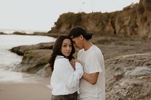 Two people share an intimate moment on a beach with rocky cliffs in the background.
