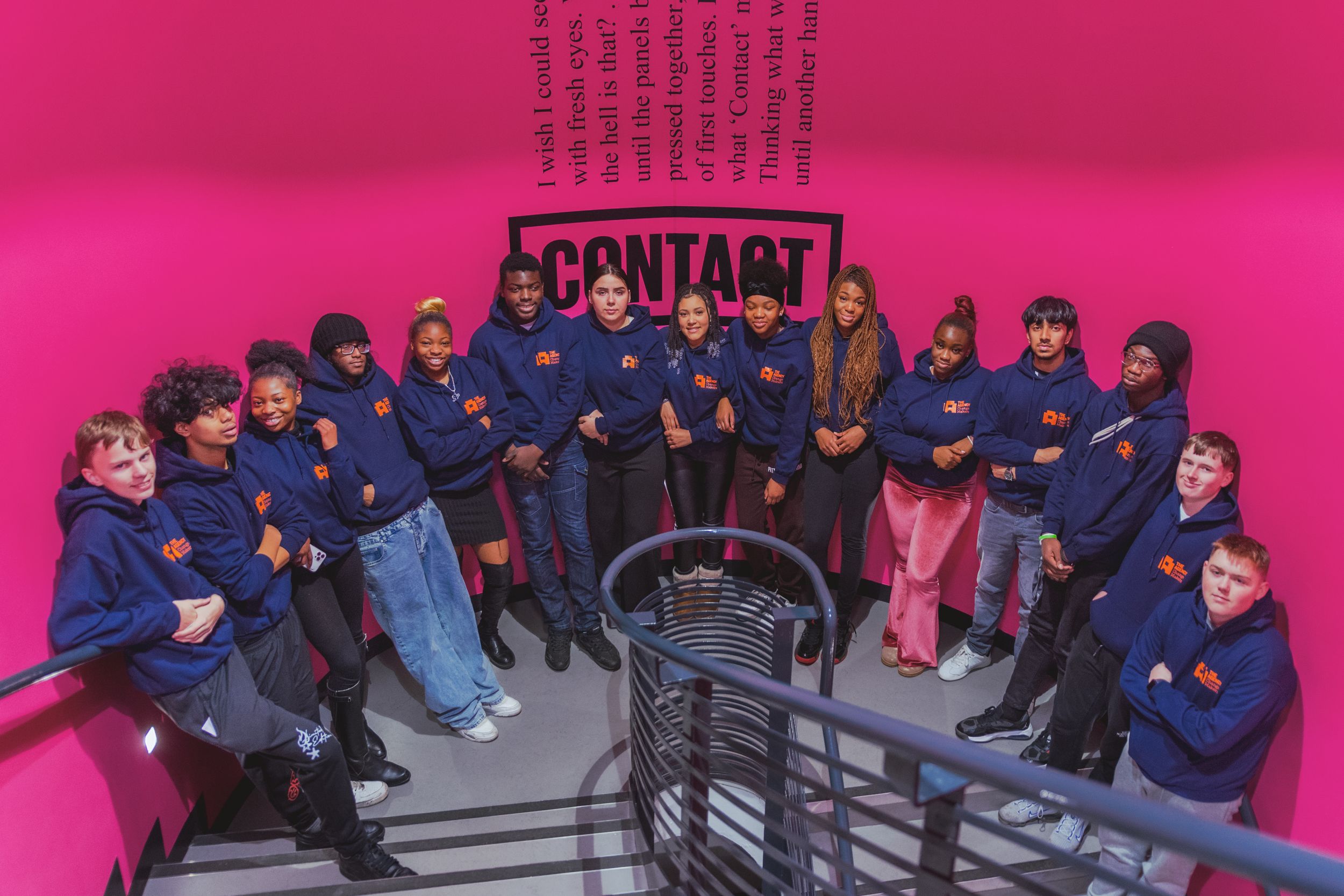 A group of young people in matching navy hoodies pose on pink stairs.