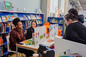 Three students collaborate at a table surrounded by books in a school library.