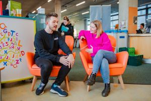Two students chat in orange chairs in a colorful modern library.