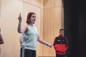 A red-haired woman gestures while leading a drama workshop with students in a wooden-paneled studio.