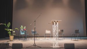 An empty stage set with a podium, microphone stand, chairs, and potted plants under soft lighting.