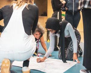 Two young women collaborate while writing on a large paper map spread on the floor.