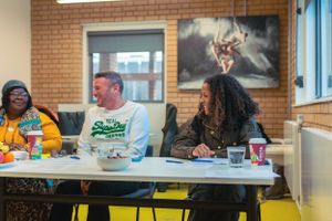 Three people laugh together at a white table in a bright community space with artwork on the wall.