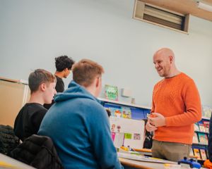 A smiling teacher in an orange sweater engages with students in a classroom.