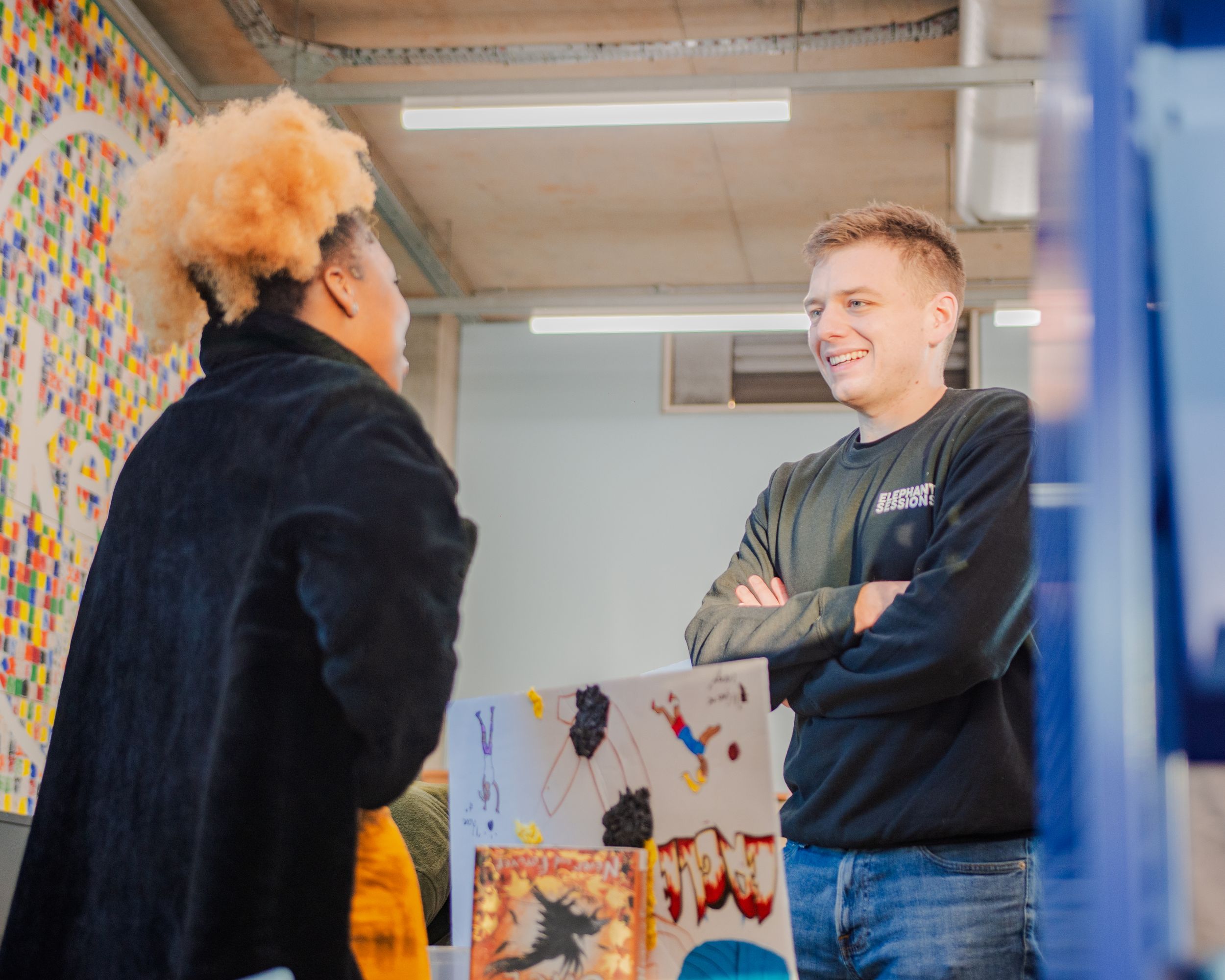 Two students smile and talk in front of colorful artwork in a studio.