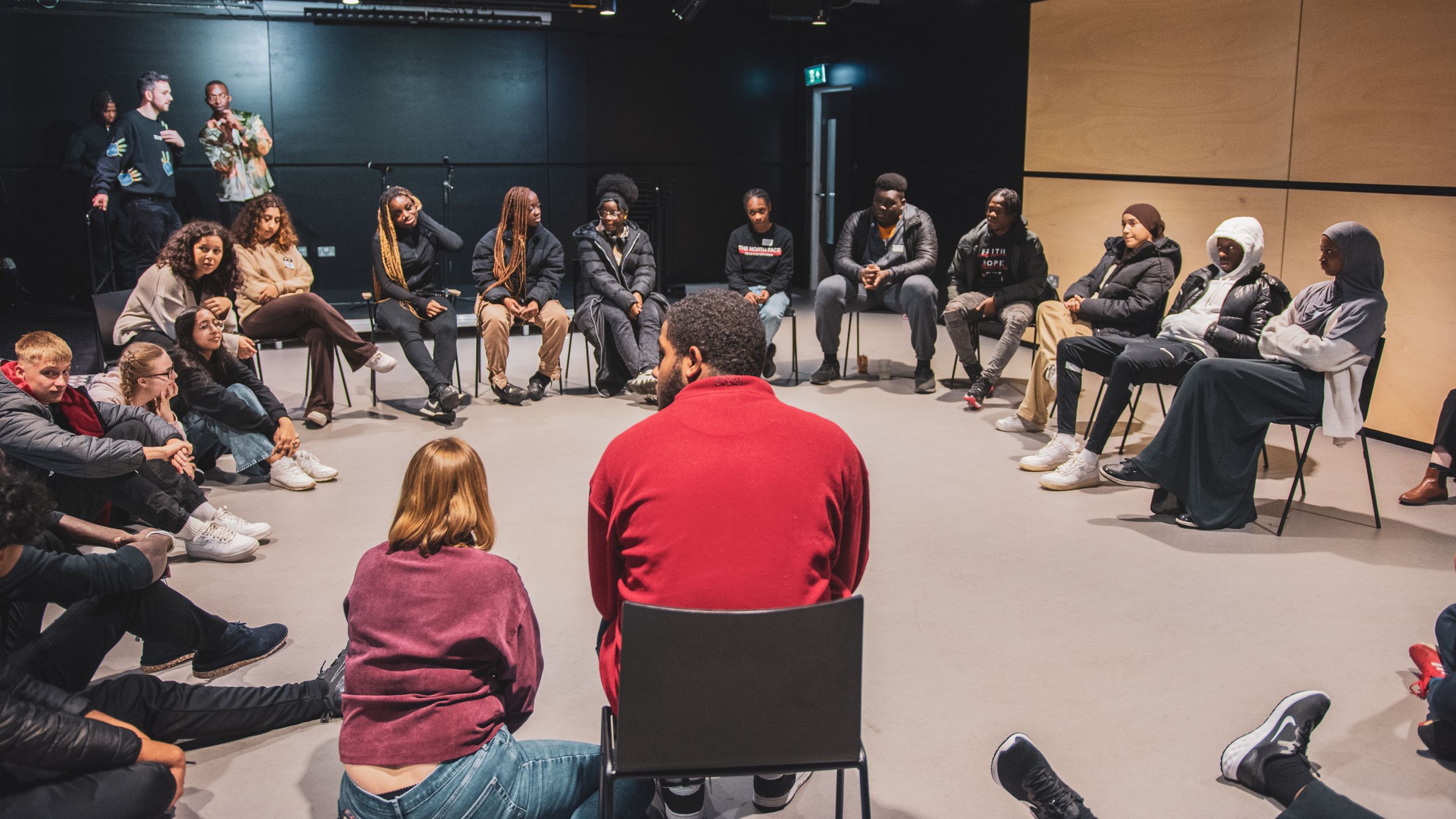 A diverse group of young people sit in a circle during a discussion workshop in a black box theater.