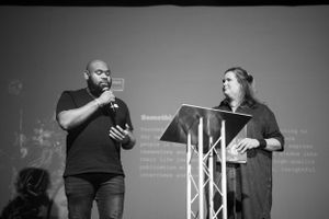 Black and white photo of a man and woman presenting together on stage with a microphone and podium.