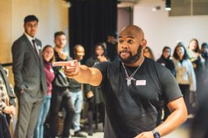 A confident man points outward while facilitating an interactive workshop with engaged participants behind him.