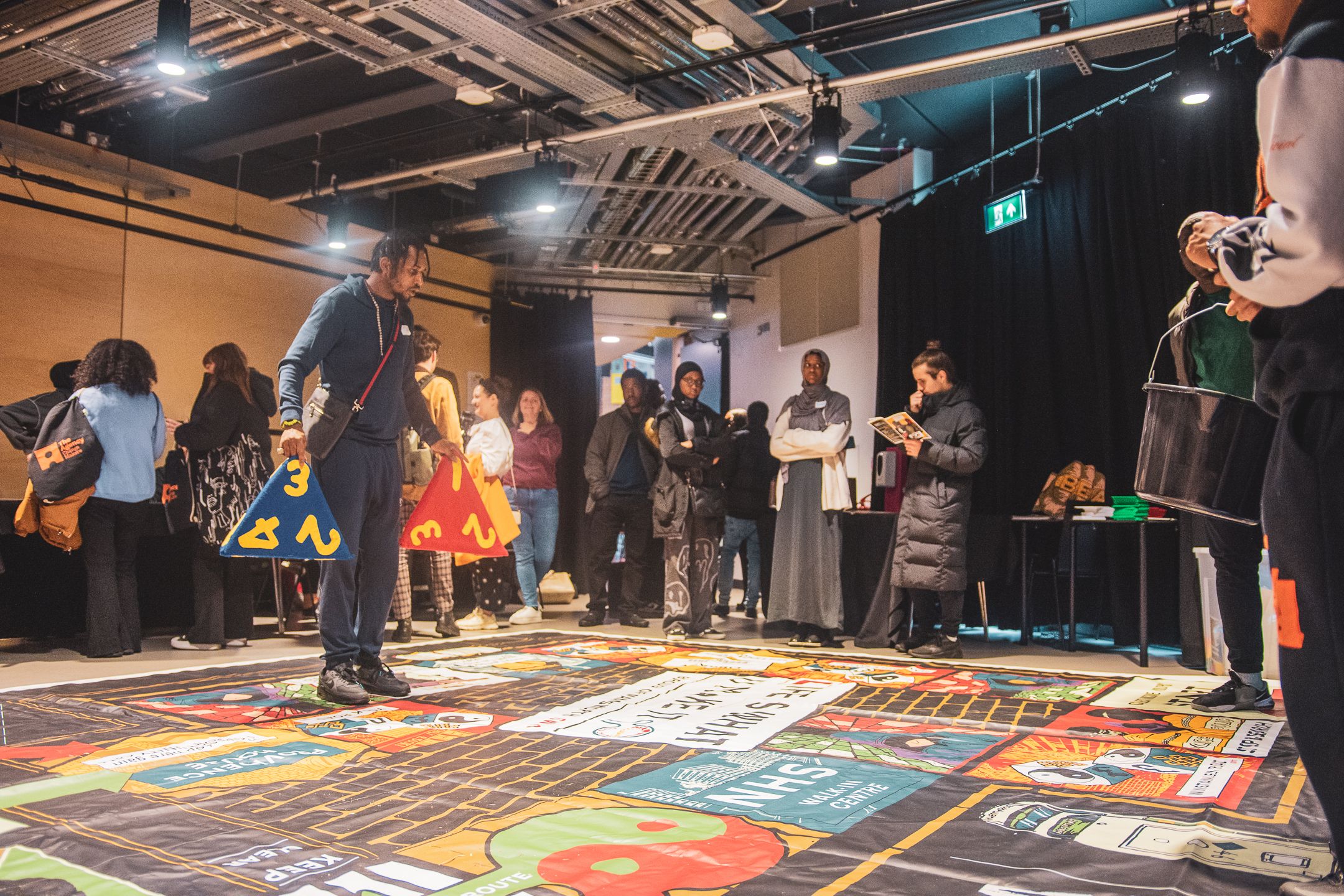 Diverse group of people gathered around a large colorful floor game installation at an indoor community event.
