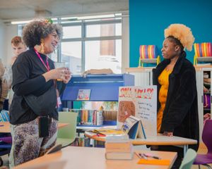 Two women conversing near a book display in a colorful library space.