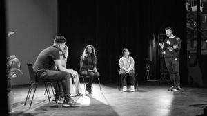 Black and white photo of four people in a drama workshop seated on chairs on a dark stage.