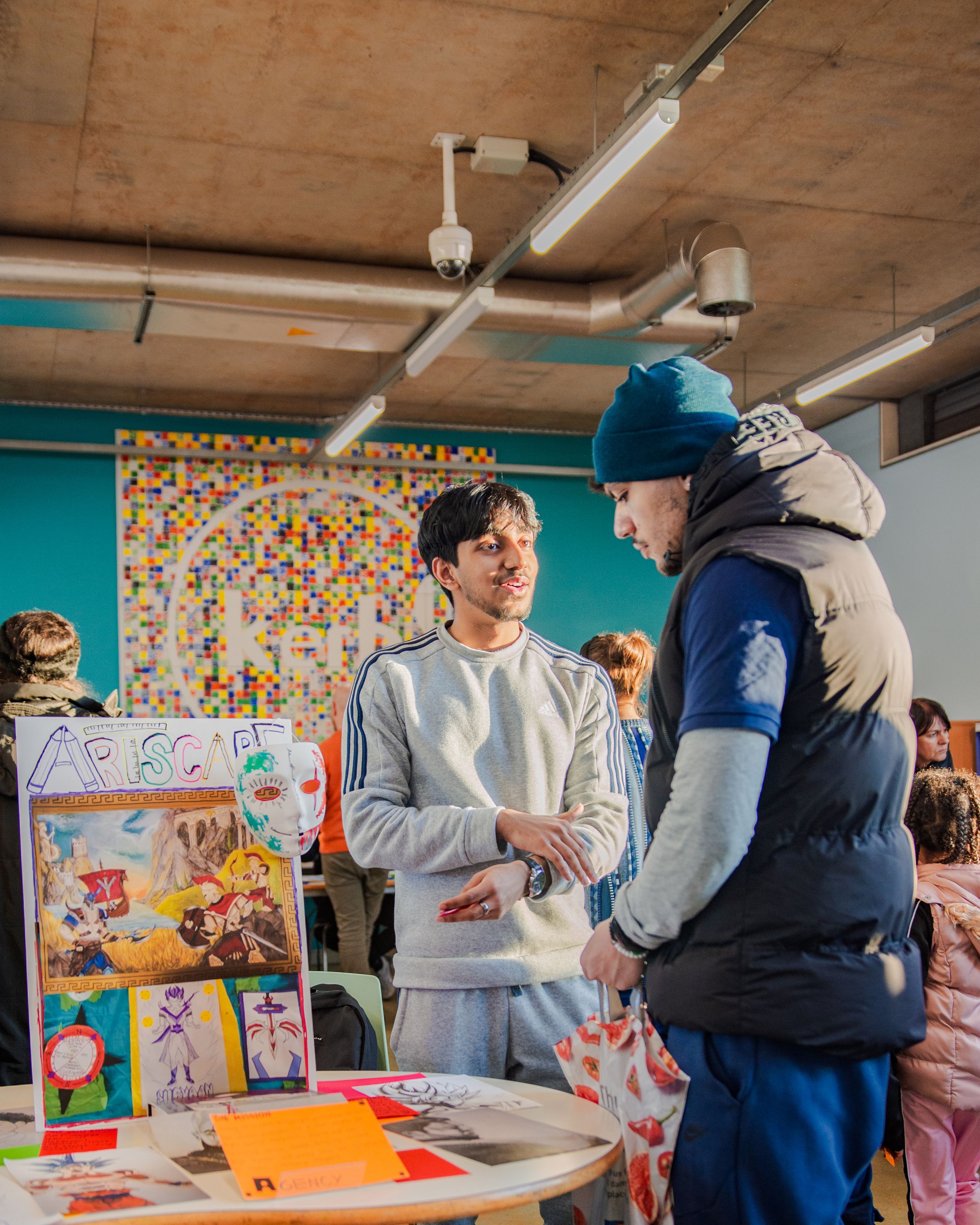 Two students discuss artwork at a creative display table in a classroom.