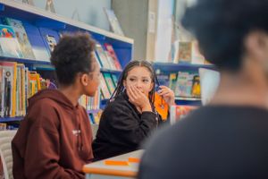 Two students sitting at a table in a library engaged in conversation.