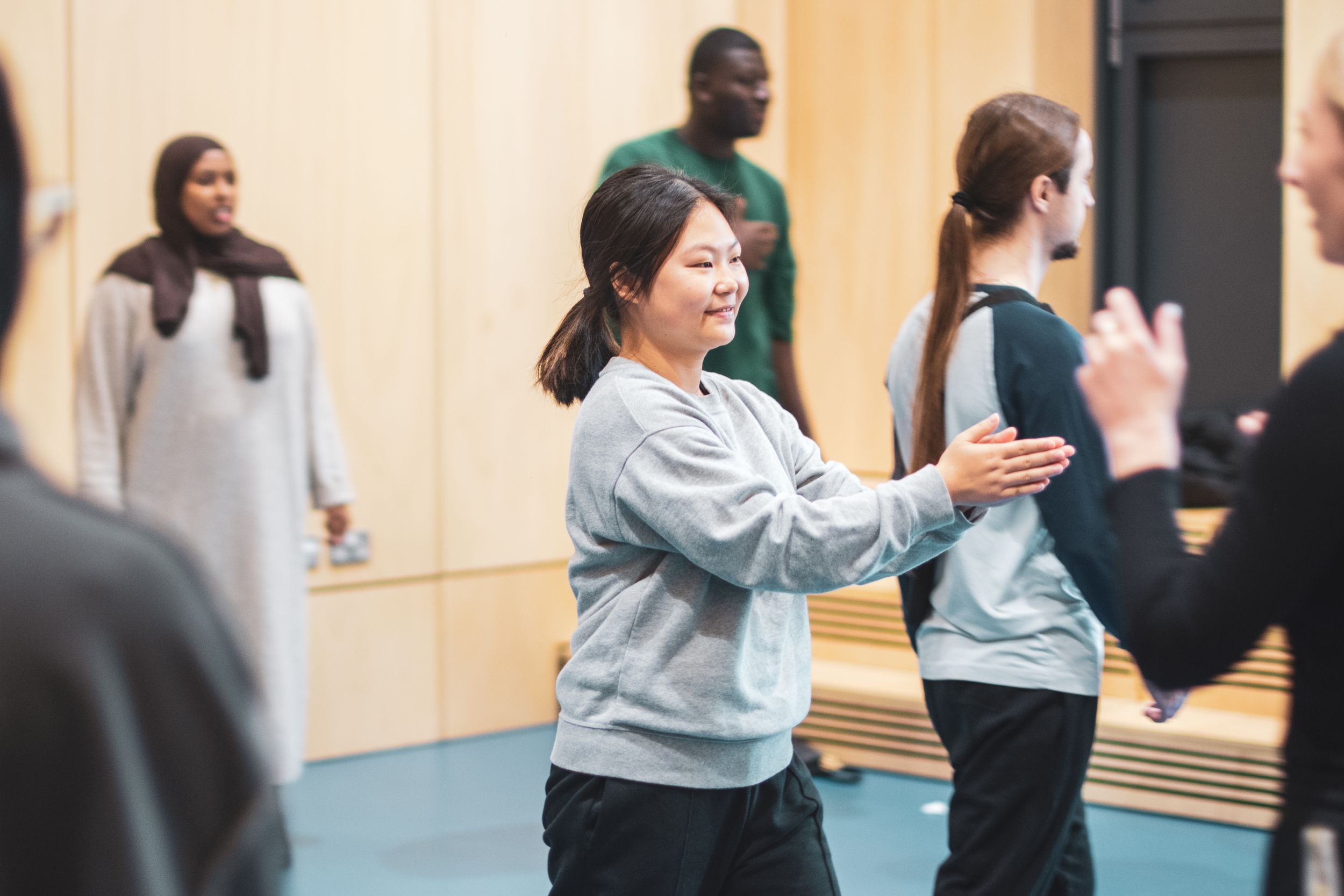 A smiling woman in a grey sweatshirt participates in a movement workshop in a mirrored studio space.