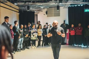 A man gestures expressively while leading a workshop for a group of young people in a black box theater.