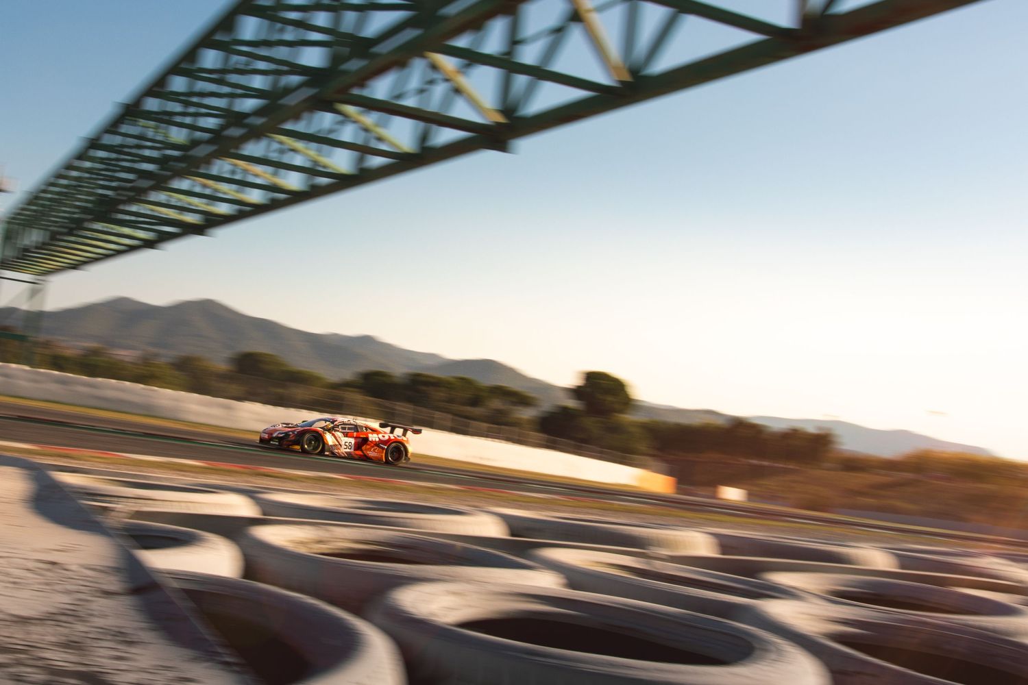 Motion-blurred race car on track under steel bridge during sunset.