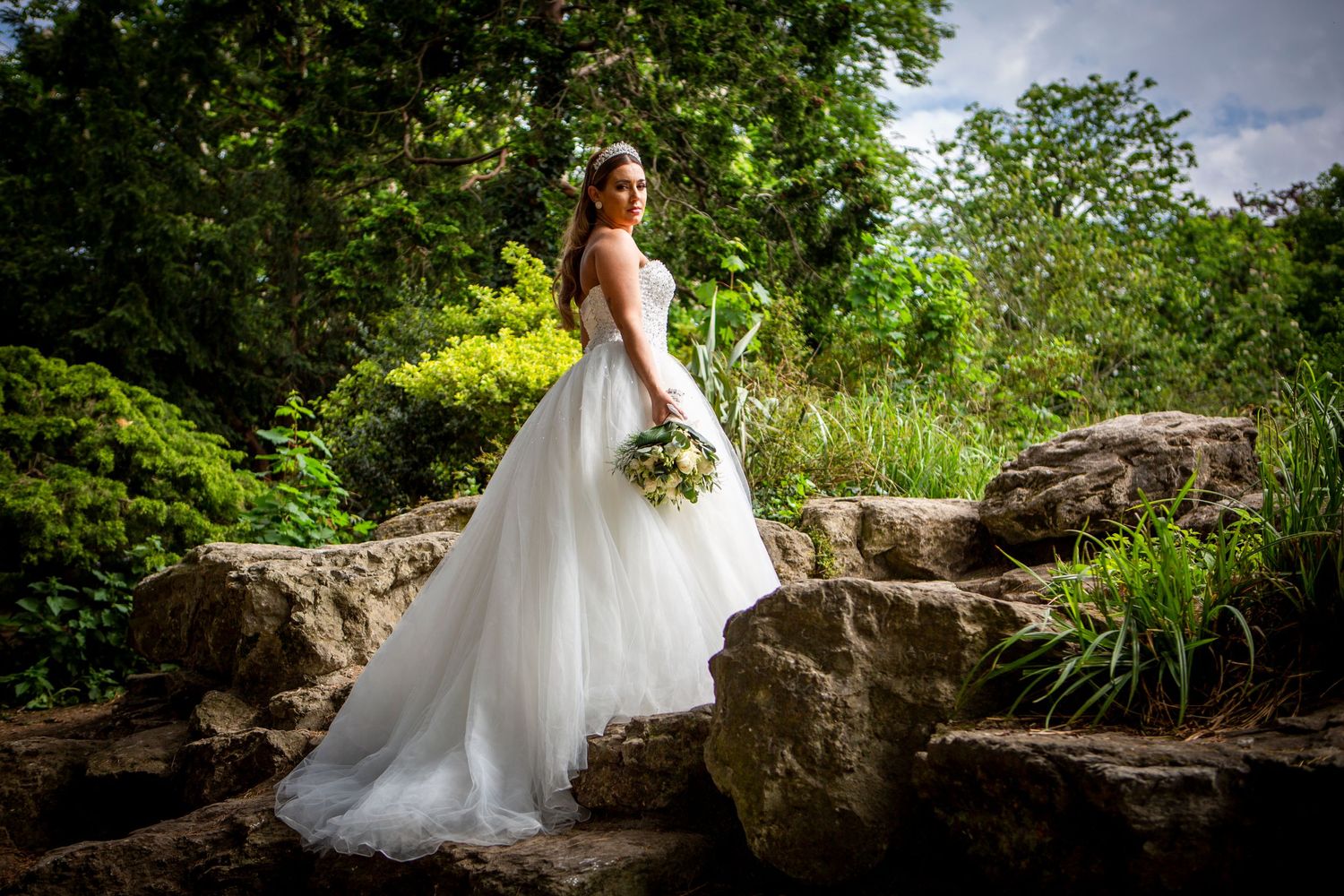 Bride in a flowing white tulle gown holds a bouquet while standing on rocks in a lush garden.