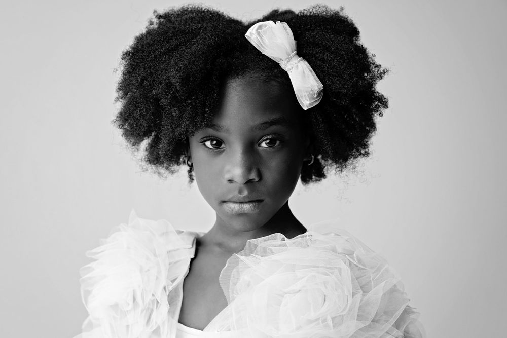 A black and white studio portrait of a young person with natural hair wearing a white bow and feathered top.
