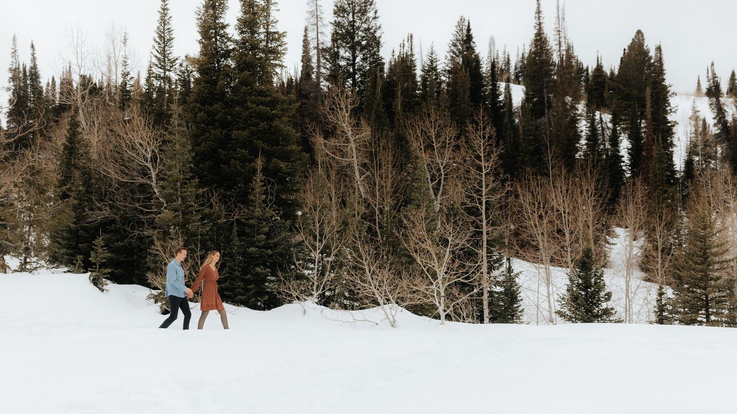 Man and woman walking along a snow covered mountain ridge with a backdrop of pine trees