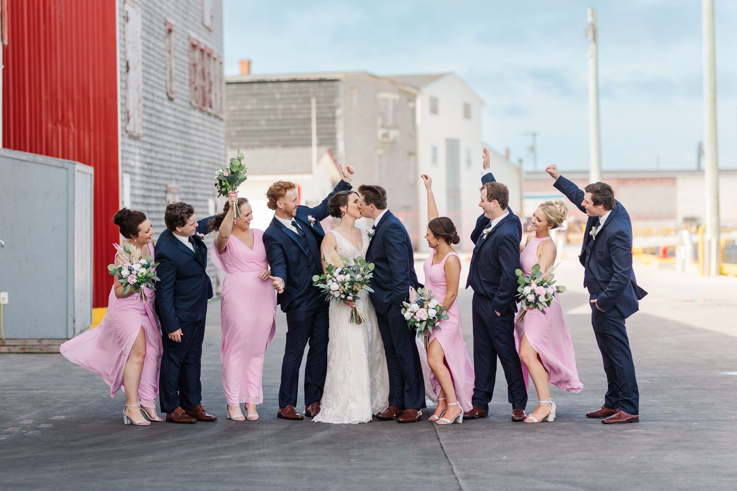 Wedding party in pink and navy attire poses together against industrial building backdrop.