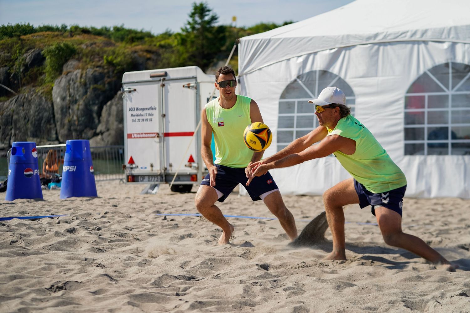 Two players dive for the ball during a beach volleyball match near a white tent on sandy shores.