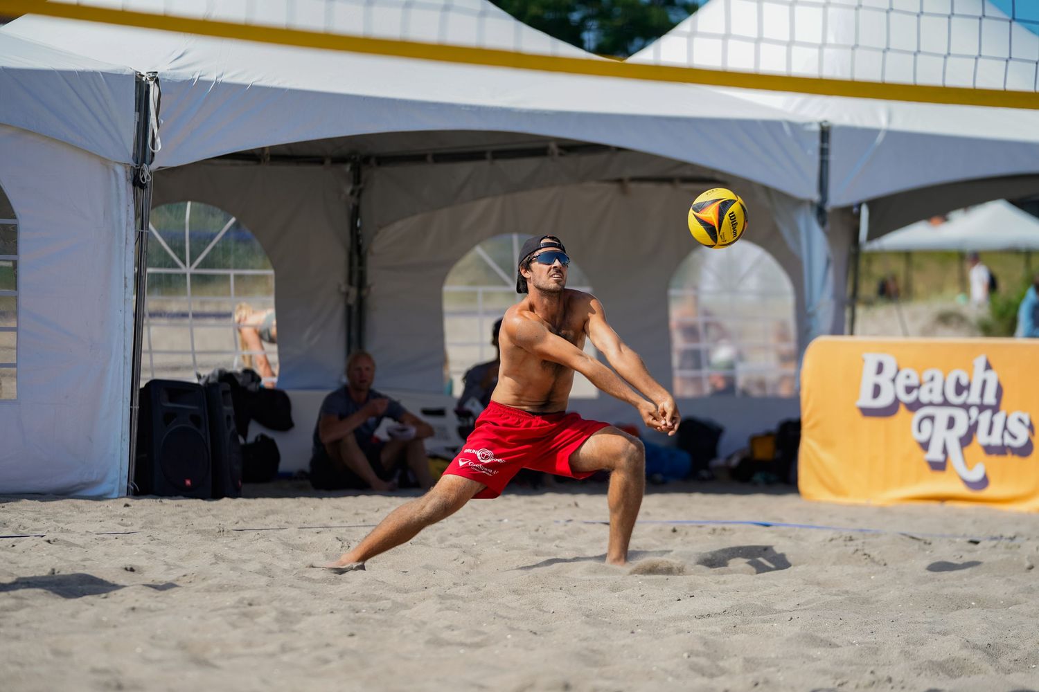 Athletic player in red shorts receives volleyball during beach tournament under tent.