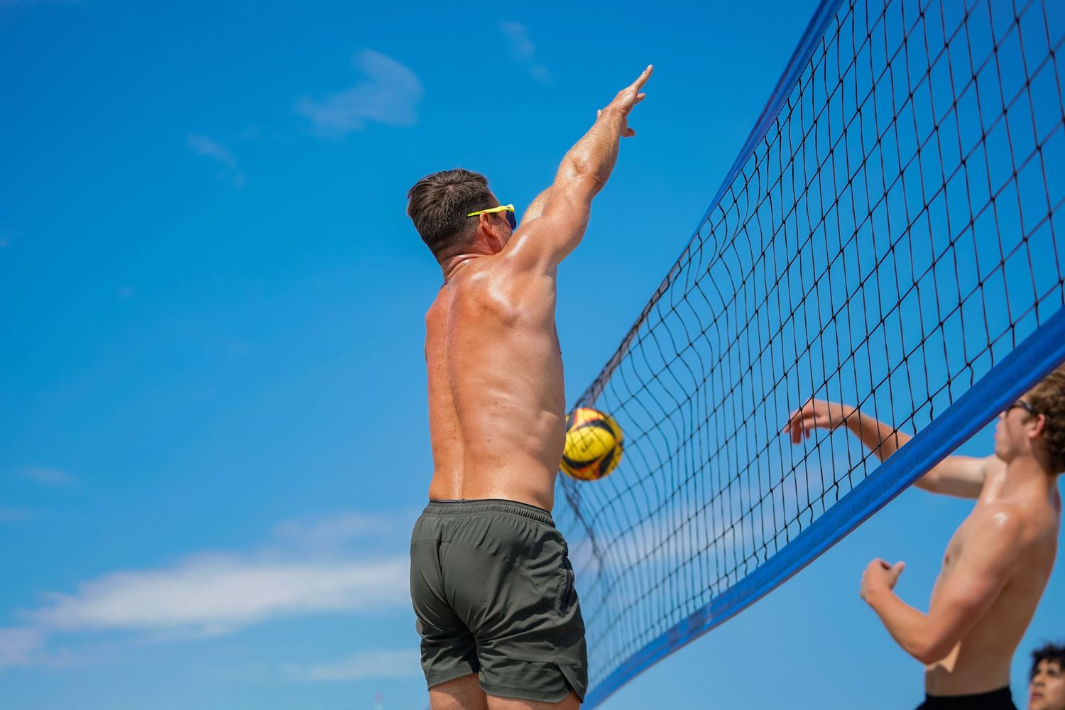Player leaps high to block a volleyball shot at the net during beach game.