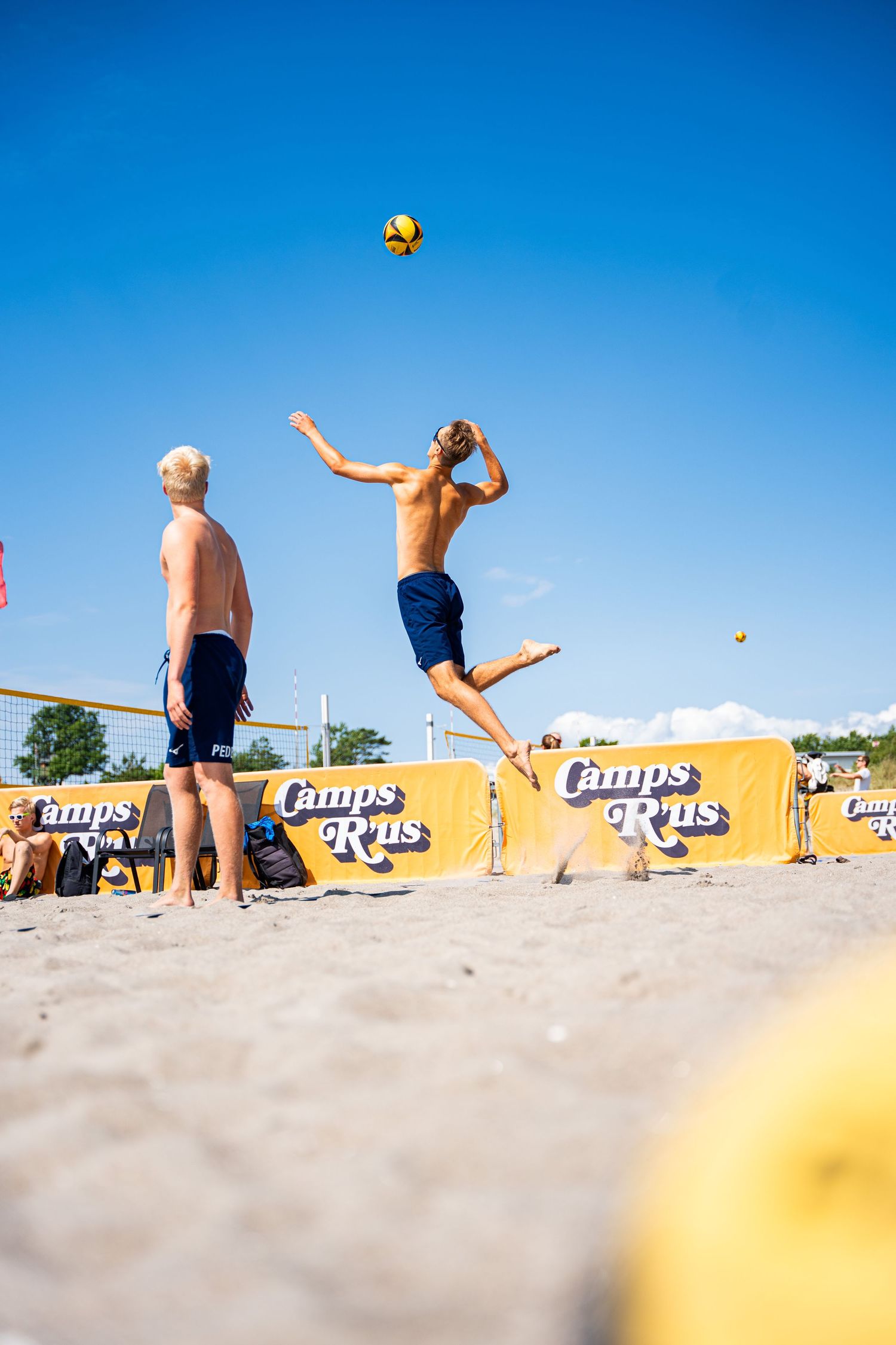 Beach volleyball player jumps to spike ball while opponent watches during sunny match.