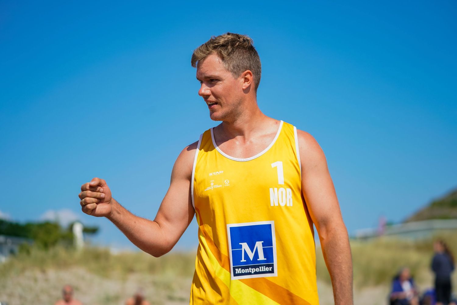 Beach volleyball player in yellow jersey stands on sandy court during tournament.