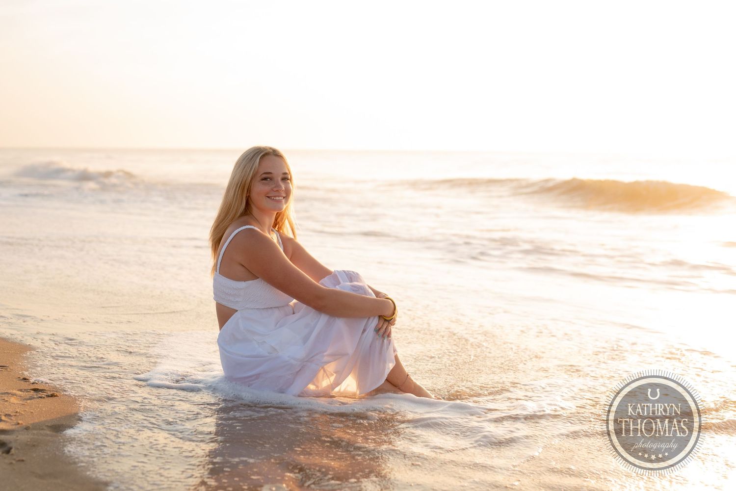 A person in a white dress sits peacefully on the beach at sunset with golden light reflecting off the ocean waves.