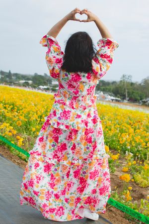 A person making a heart shape with their hands while wearing a floral dress in a flower field.