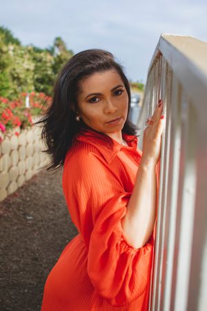 Someone in a vibrant orange dress poses elegantly against a white railing outdoors.