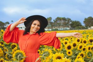 Dramatic fashion pose in a sunflower field wearing a bright orange dress against blue sky.