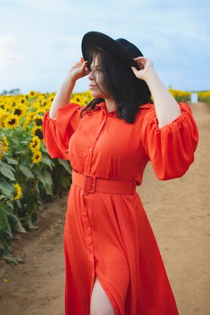 A bright orange maxi dress with puff sleeves and black hat stands in front of vibrant sunflower field.