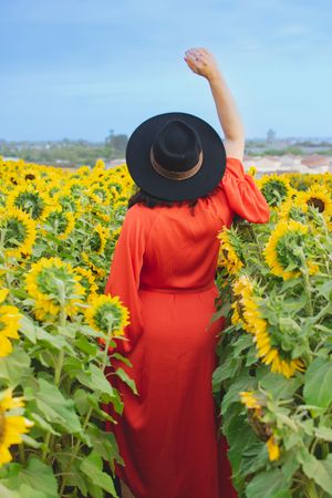 Back view of figure in flowing red dress raising arm in sunflower field against blue sky.