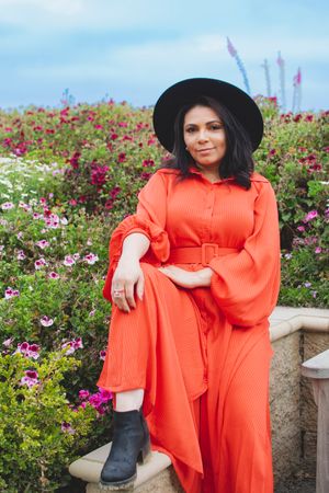Figure in orange dress and black hat poses on garden bench surrounded by colorful flowers.