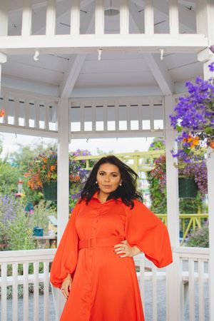 Figure in bright orange dress poses in white gazebo surrounded by hanging flower baskets.