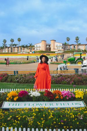 A bright orange dress against colorful flower fields with palm trees and buildings in the background.