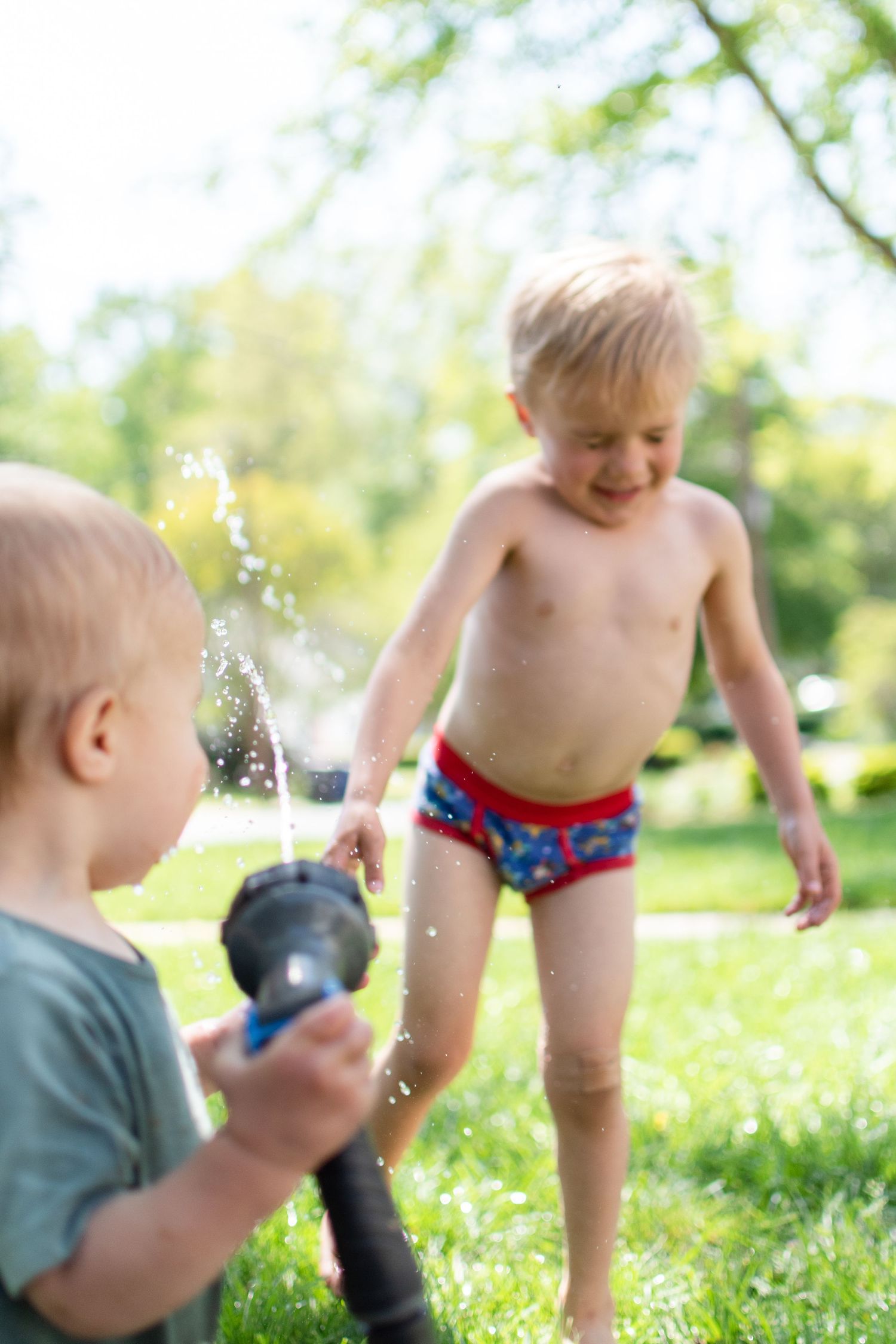 Baby bother squirts the water hose at his big brother in his front yard in the summer.