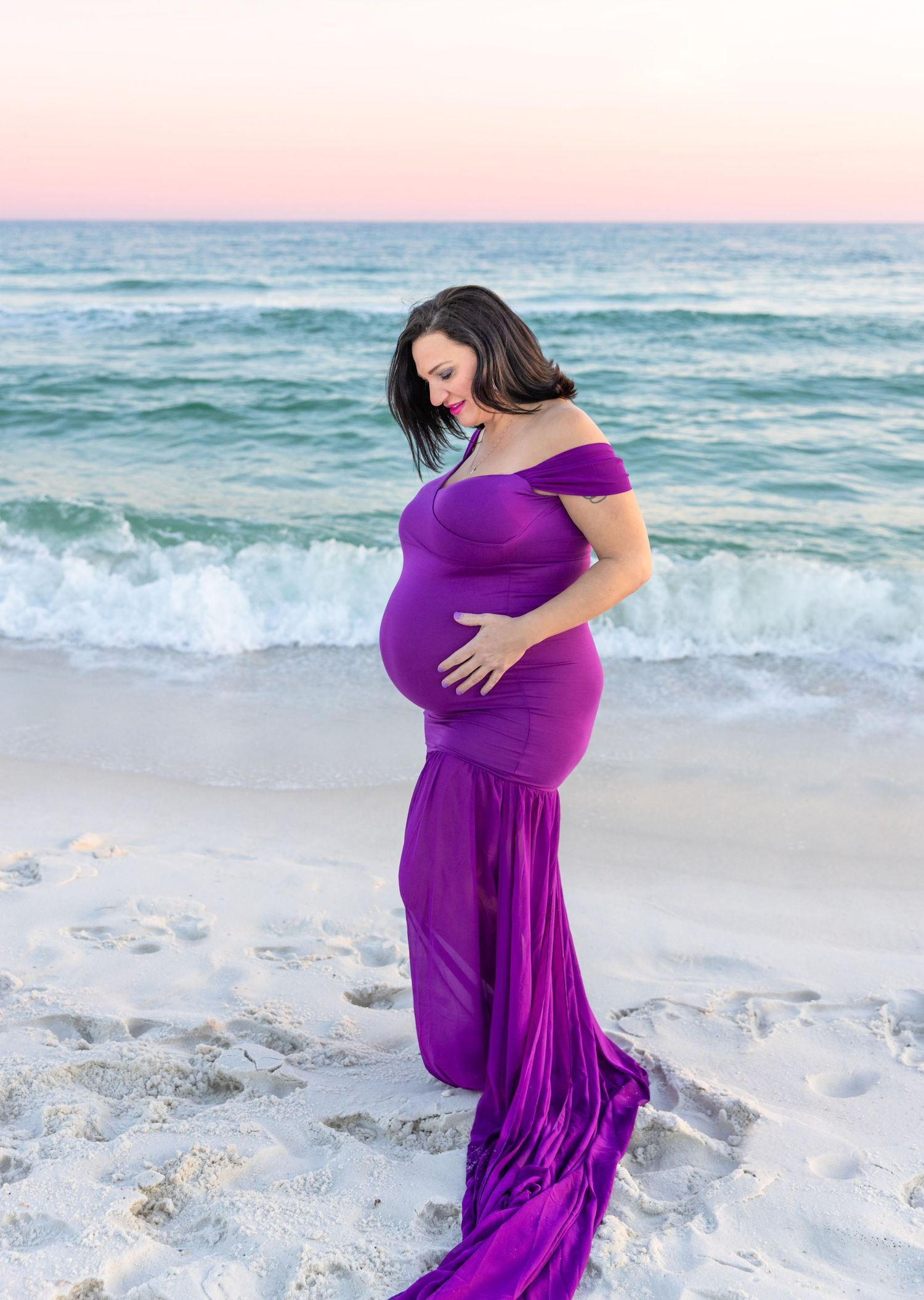 Maternity photo of woman in flowing purple dress on beach at sunset.