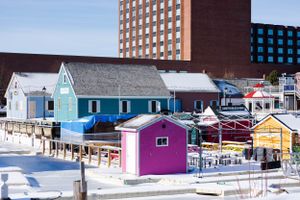 Colorful fishing shacks and buildings along snowy waterfront with apartment building in background.